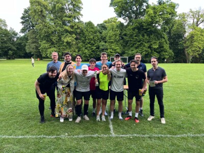 Ein Team in weinroten Trikots posiert mit einem Pokal und ihren Fans für ein Gruppenfoto. Die Stimmung ist ausgelassen, im Hintergrund ein Fußballtor auf dem Rasen.