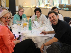 Mehrere Teilnehmende sitzen an einem gedeckten Tisch, lachen und unterhalten sich in entspannter Atmosphäre.