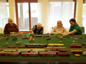 Vier Personen, darunter drei ältere Menschen und eine Betreuerin, sitzen um eine große Modelleisenbahnanlage auf einem Tisch. Die Szene zeigt verschiedene Züge, Gebäude und Landschaftselemente. Eine Seniorin greift lachend nach einem Zug, während die Betreuerin lächelnd mitmacht. Die Atmosphäre wirkt fröhlich und gemeinschaftlich.