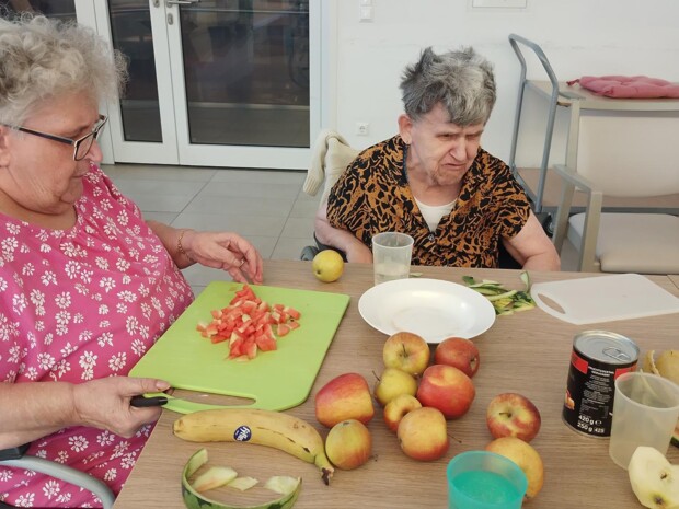 Zwei Bewohnerinnen sitzen an einem Tisch und bereiten Obst vor. Eine Bewohnerin schneidet Wassermelone auf einem grünen Brett, während Äpfel, Bananen, Melonenschalen und weitere Zutaten auf dem Tisch liegen.