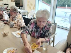 Eine ältere Frau löffelt Mais auf ein rechteckiges Stück Pizzateig. Weitere vorbereitete Teige liegen daneben auf Backpapier. Rundherum sitzen weitere Senior:innen, die ebenfalls beim Belegen ihrer Pizzen sind. Im Hintergrund ist ein heller Aufenthaltsraum mit großen Fenstern zu sehen.
