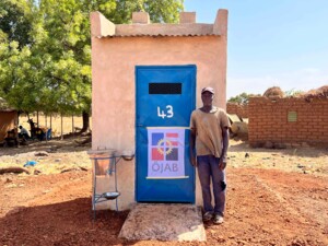 Eine Person steht vor einem kleinen, freistehenden Gebäude mit einer blauen Tür, die die Nummer 43 trägt und mit dem Logo der ÖJAB gekennzeichnet ist. Neben dem Eingang befindet sich eine einfache Handwaschanlage. Im Hintergrund sind Bäume, Lehmhäuser und sitzende Personen im Schatten zu sehen, was ein klassisches, ländliches Dorfumfeld darstellt.
