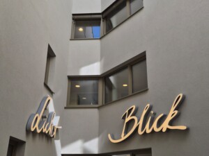Innenhof eines modernen Gebäudes mit mehreren Fensterfronten. An den hellen Wänden sind in geschwungener Leuchtschrift die Wörter "Licht" und "Blick" angebracht.
