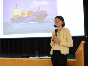 Eine Vortragende steht mit Mikrofon vor einer Präsentationsfolie mit einem Containerschiff und erklärt Inhalte im Veranstaltungssaal.