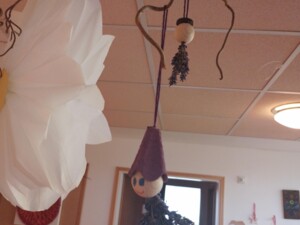 Ein Lavendelwichtel, hergestellt aus Lavendelzweigen, einer Holzkugel als Kopf und einer lilafarbenen Mütze, hängt zusammen mit einem großen Papierblumen-Dekorationselement an einem dekorativen Zweig von der Decke eines Gemeinschaftsraums.