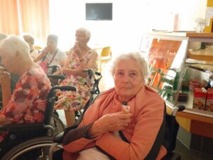 Eine Bewohnerin sitzt im Rollstuhl und hält ein Eis in der Hand. Im Hintergrund essen weitere Bewohner:innen Eis; daneben befindet sich eine kleine Bar mit Getränken.