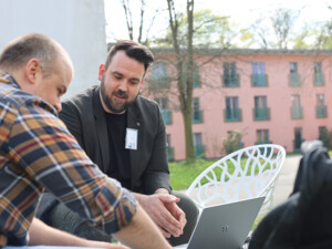 Zwei Personen sitzen draußen an einem Tisch, arbeiten gemeinsam an einem Laptop und besprechen Inhalte.