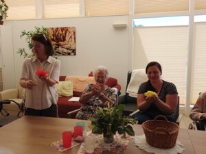 Eine Begleitperson steht im Gemeinschaftsraum und hält ein rotes Tuch. Drei sitzende Teilnehmer:innen halten farbige Tücher oder Gegenstände in den Händen. Auf dem Tisch stehen ein Korb, Blumen, Becher und Servietten.