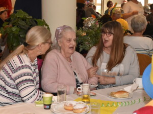 Drei Frauen sitzen eng beisammen an einem Tisch. Eine ältere Frau in rosa Jacke singt mit geöffnetem Mund, während zwei jüngere Frauen neben ihr sie begleiten und unterstützen. Auf dem Tisch stehen Getränke und Gebäck. Im Hintergrund sind weitere Gäste des Festes zu sehen.