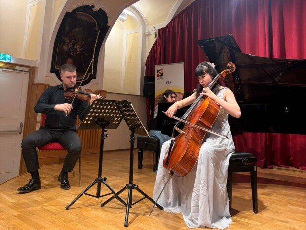 Zwei Musiker:innen spielen gemeinsam Violine und Violoncello. Sie sitzen auf der Bühne mit Notenständern vor sich. Im Hintergrund ist ein Flügel zu sehen an dem eine dritte Musikerin sitzt, der Raum ist warm beleuchtet und von einem roten Vorhang eingerahmt.