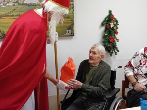 Ein älterer Mensch im Rollstuhl bekommt von einer als Nikolaus verkleideten Person ein rotes Geschenk überreicht. Im Hintergrund ist weihnachtlicher Wandschmuck zu sehen.