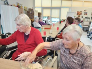 Zwei ältere Personen sitzen an einem Tisch und panieren gemeinsam Pilzstücke in einer Schüssel mit Bröseln. Im Hintergrund sind weitere Bewohner:innen in einer Gruppe zu sehen.