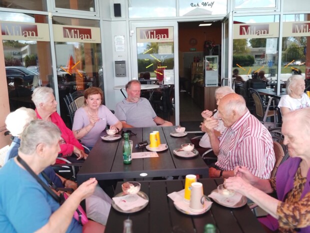 Eine Gruppe Senior:innen sitzt gemeinsam an zwei Tischen vor dem Café Milan. Alle essen Eis und unterhalten sich. Die Stimmung wirkt fröhlich und gemeinschaftlich.