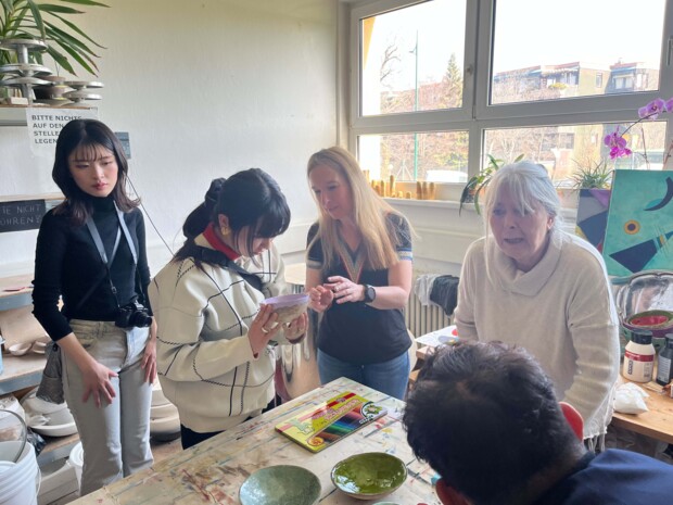 Vier Personen stehen in einem lichtdurchfluteten Atelier an einem mit Farben und Schalen bedeckten Arbeitstisch. Zwei ältere Anleitende erklären etwas, während zwei jüngere Personen aufmerksam zuhören und eine davon eines der Stücke in den Händen betrachtet. Auf dem Tisch liegen bunte Stifte, Schalen mit grüner Glasur oder Farbe sowie weitere Kreativmaterialien. Im Hintergrund sorgen große Fenster, Pflanzen und ein Gemälde für eine freundliche Workshop-Atmosphäre.