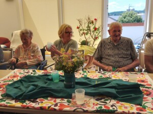 Mehrere Bewohner:innen und Begleiter:innen sitzen an einem gedeckten Tisch mit Blumenstrauß und bunten Stoffen. Eine Person hält ein Tuch in der Hand, während andere aufmerksam zuhören und mitmachen.
