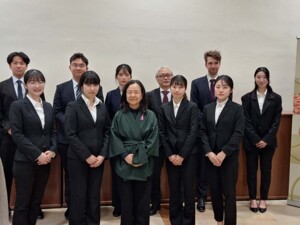 Eine Gruppe formell gekleideter, meist junger Menschen posiert in zwei Reihen für ein Foto. Auf der rechten Seite des Bildes steht ein Roll-up mit der Aufschrift "Botschaft Japan Österreich" und einigen bunten, traditionell ostasiatischen Motiven.