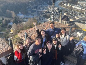 Acht Personen posieren lächelnd auf einer hölzernen Aussichtsplattform vor einer weitläufigen Stadtlandschaft. Einige winken oder zeigen ein Victory-Zeichen. Im Hintergrund sind zahlreiche historische Gebäude, Kirchen, ein Fluss und bewaldete Hügel zu sehen. Das Wetter ist klar.