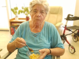 Eine ältere Person sitzt auf einem Stuhl und isst mit einem Löffel aus einer kleinen Glasschüssel frischen Obstsalat. Im Hintergrund ist ein Rollator zu sehen. Die Atmosphäre wirkt hell und freundlich.