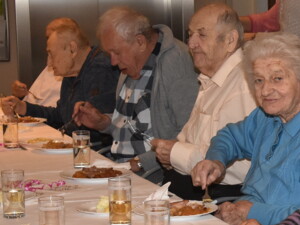 Eine Gruppe älterer Menschen sitzt an einem langen Tisch und isst gemeinsam. Die Teller sind mit traditionellen Gerichten wie Sauerkraut, Knödeln und Fleisch befüllt.