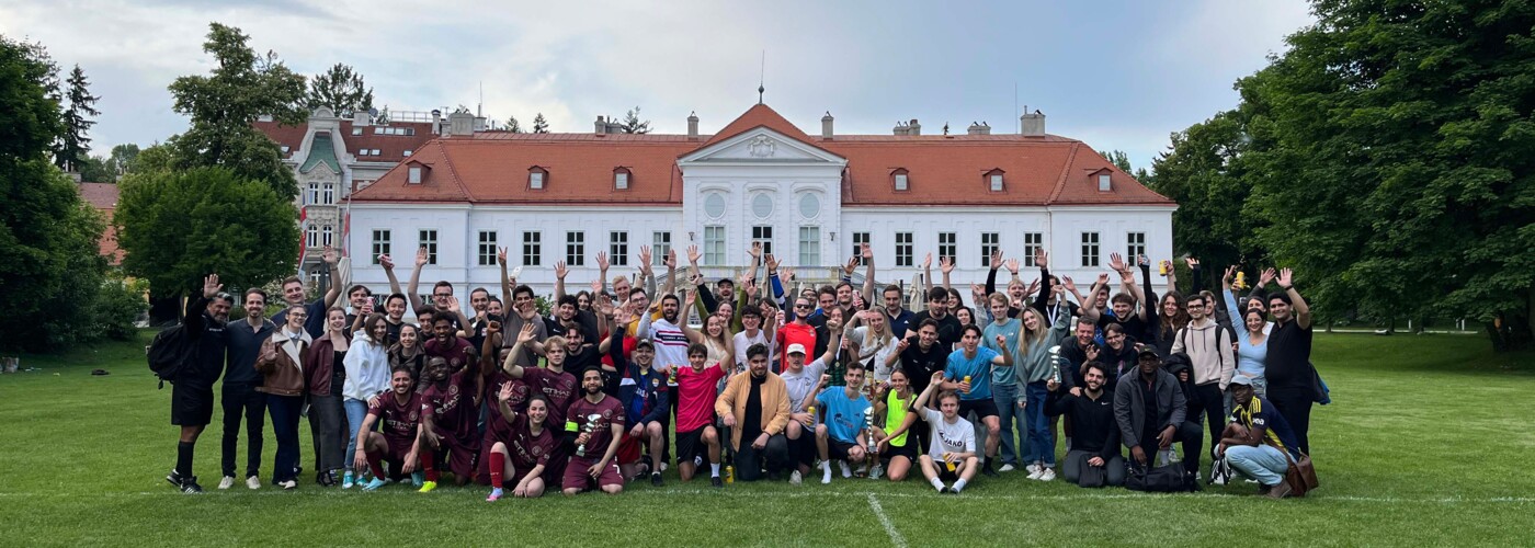 Eine große Gruppe von Menschen, darunter Spieler:innen und Zuschauer:innen, posiert lachend und mit erhobenen Armen auf einer Wiese. Im Hintergrund das weiße Gebäude des Europahauses mit rotem Dach und Bäumen. 