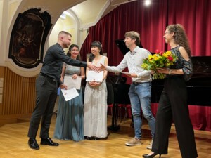 Mehrere junge Musiker:innen stehen auf einer Bühne vor rotem Vorhang und einem Flügel. Eine Person überreicht einer anderen die Hand und Urkunden, während zwei weitere Musiker:innen daneben stehen und lächeln. Eine Person hält einen Blumenstrauß. Die Szene zeigt die feierliche Auszeichnung nach einem Konzert.