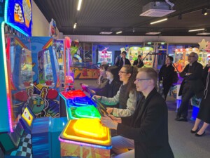 In einem bunt leuchtenden Innenraum spielen vier junge Menschen ein Arcade-Spiel. Die Spieler:innen sind von der Seite fotografiert und spielen an je einem Lenkrad ein Renn-Videospiel. Hinter ihnen am Bildrand sind weitere Personen, die das Spiel amüsiert betrachten, zu sehen. Im Hintergrund sind weitere Arcade-Spiele, etwa Kranspiele oder ein Airhockey-Tisch.