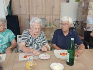 Drei ältere Personen sitzen nebeneinander am Tisch und belegen ihre Pizzateige. Eine Person greift nach Schinken, eine andere hat bereits Tomatensauce aufgetragen. Auf dem Tisch stehen eine große Wasserflasche sowie Schälchen mit Mais, Schinken und Käse.