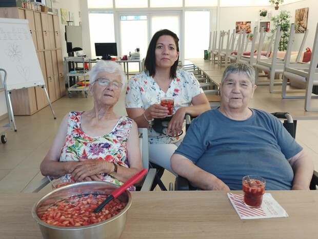 Zwei ältere Bewohner:innen sitzen mit einer Senior:innenbetreuerin an einem Tisch in einem hellen Gemeinschaftsraum. Vor ihnen steht eine große Schüssel mit roter Sommerbowle und Fruchtstücken, daneben Gläser mit dem Getränk.
