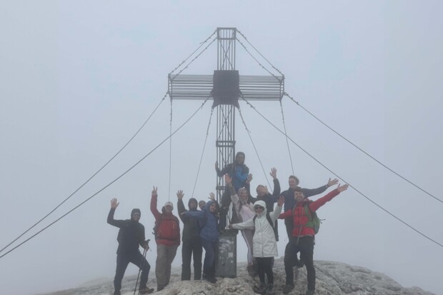 Zehn Personen jubeln vor einem großen Gipfelkreuz aus Metall im Nebel. Alle haben die Hände in die Höhe gestreckt. Das Wetter ist neblig, die Sicht eingeschränkt, die Stimmung aber sichtbar ausgelassen.