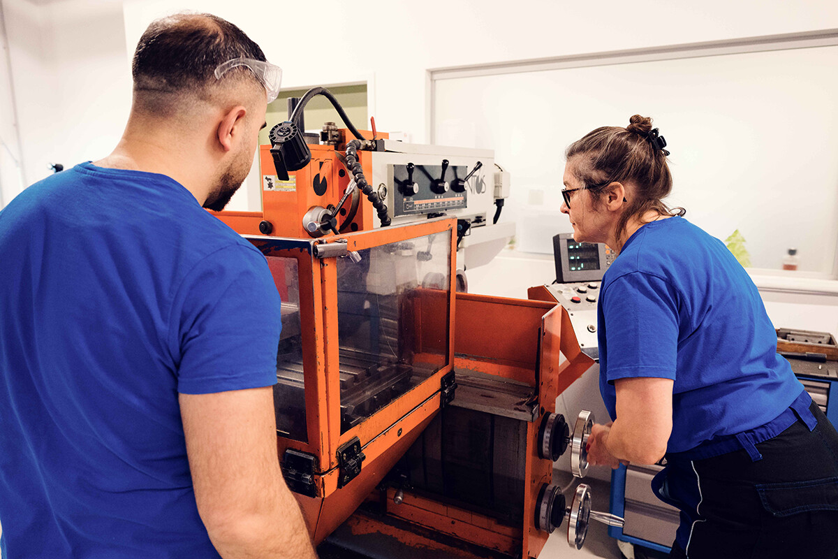 Zwei Kursteilnehmende in blauen Shirts arbeiten an einer großen orangefarbenen Industriemaschine. Eine Person steht links vor der Maschine, eine weitere bedient rechts ein Handrad und blickt auf die Steuerung. Foto: www.inframe.at