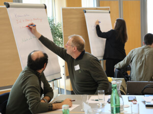 Zwei Gruppen von Teilnehmenden schreiben Stichworte auf Flipcharts, während andere aufmerksam zuhören und Ideen einbringen.