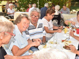 Mehrere Personen sitzen eng beisammen an einem langen Tisch im Garten. Sie essen, trinken und unterhalten sich. Eine Frau in der Mitte lacht herzlich, auf den Tellern liegen Fleisch, Salat und Beilagen.