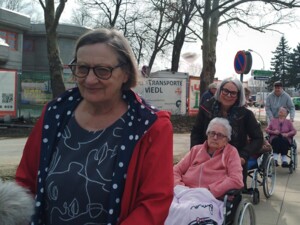 Mehrere Bewohner:innen sitzen in Rollstühlen und werden von Begleitpersonen auf einem breiten Weg im Freien geschoben. Die Gruppe ist bei sonnigem Wetter in einer Reihe unterwegs, links und rechts sind Wiese, Gebäude und parkende Autos zu sehen.