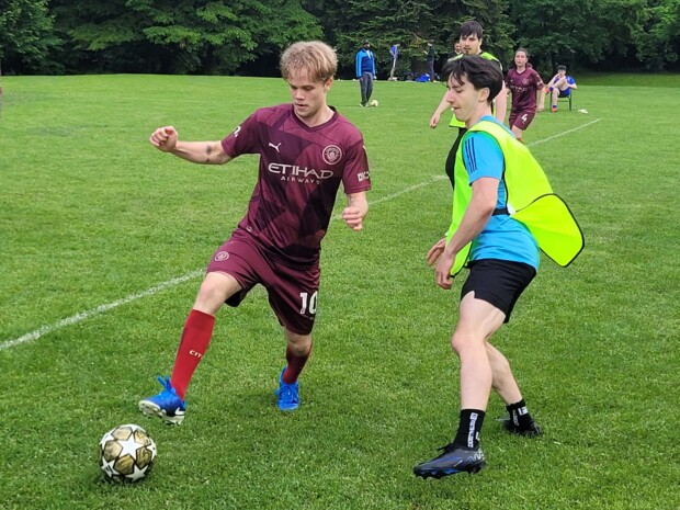 Zwei Fußballspieler liefern sich ein intensives Duell um den Ball. Eine Person trägt ein weinrotes Trikot, die andere ein gelbes Leibchen über blauem Shirt. Im Hintergrund sind weitere Spieler:innen und Zuschauer:innen zu sehen. 