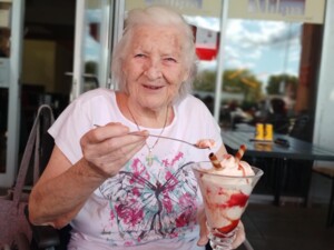 Eine Seniorin im Rollstuhl hält einen großen Eisbecher mit Erdbeersoße, Sahne und Schokoröllchen. Sie lächelt herzlich und wirkt sichtlich erfreut.