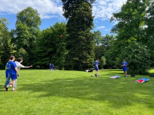 Personen werfen bunte Säckchen bei einem Cornhole-Spiel auf Rasen. Einige tragen Fußballtrikots, im Hintergrund stehen weitere Mitspielende. Die Szene spielt sich in einem von Bäumen umgebenen Bereich ab. 