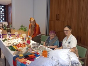 Mehrere Bewohner:innen und eine Betreuungsperson sitzen und stehen hinter einem Tisch mit handgefertigten Produkten wie gestrickten Decken, Polstern und Bastelarbeiten. Die Beteiligten wirken zufrieden und engagiert, während sie ihre Waren präsentieren. Die Umgebung ist hell und freundlich und unterstreicht die gemeinschaftliche Stimmung.