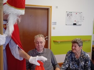 Ein als Nikolaus verkleideter Mensch überreicht einem älteren Bewohner im Rollstuhl ein rotes Geschenksäckchen. Neben dem Bewohner sitzt eine weitere Person, die seine Hand hält. Die Szene spielt sich an einem Tisch im Pflegeheim ab.