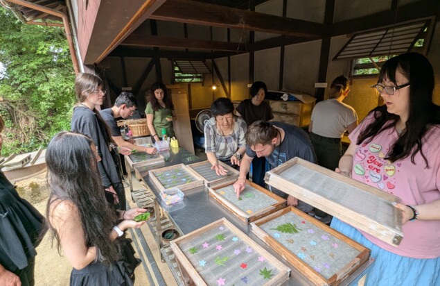 Eine Werkstatt, die links ins Freie geöffnet ist. Auf einem Metalltisch liegen mehrere Rahmen mit japanischem Papier, Blättern und ausgeschnittenen Papierformen. Junge europäische Reisende arbeiten unter Anleitung einer älteren Japanerin an dem Papier. Eine Teilnehmerin hält einen dieser Rahmen, andere dekorieren eigenes in Rahmen gespanntes Papier. Im Hintergrund sehen vereinzelt Personen zu oder arbeiten selbst daran. Das Innere der Werkstatt zeigt eine traditionell japanische Architektur. Draußen sind grüne Pflanzen zu sehen. Foto: Zoltan Hajduk