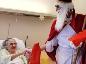 Eine ältere Person im Nachthemd liegt im Bett und greift freudig nach der Hand des Nikolaus, der ein Geschenk überreicht. Ein gelber Becher steht auf dem Nachttisch.