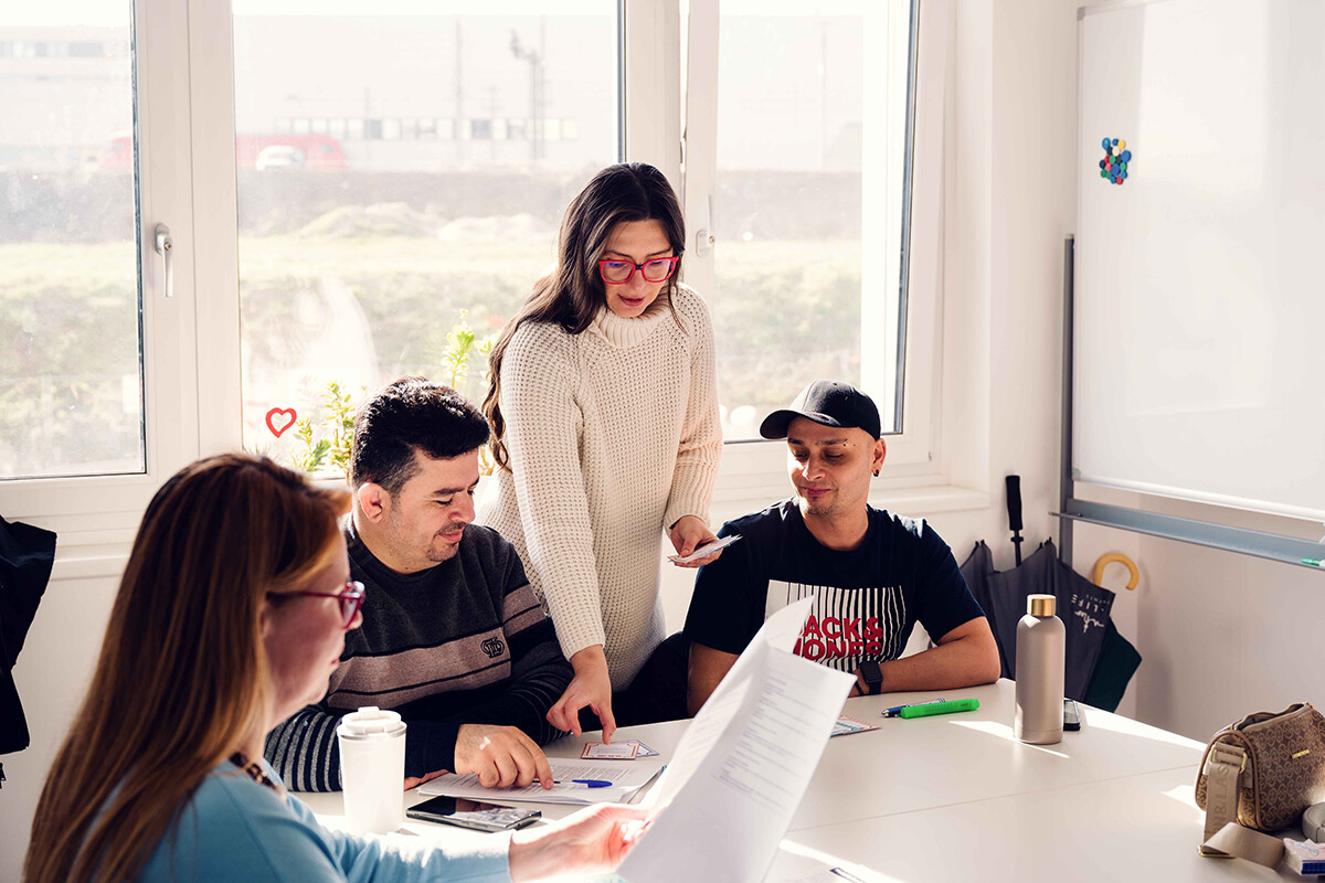 Eine Kursleiterin steht an einem Tisch mit mehreren Kursteilnehmenden und zeigt auf ein Arbeitsblatt. Die Gruppe sitzt in einem hellen Raum zusammen und arbeitet mit ausgedruckten Unterlagen. Foto: www.inframe.at