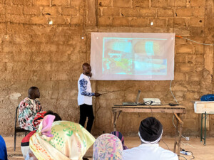 Ein burkinischer Mann steht in einem einfachen Schulungsraum und hält einen Vortrag. Er zeigt auf eine an die Wand projizierte Präsentation mit Bildern von Bienenstöcken, während mehrere Teilnehmende, darunter Frauen mit Kopfbedeckungen, auf Bänken sitzen und zuhören. Vor ihm stehen ein Tisch mit Laptop und Beamer; die Lehmwand des Raumes und die schlichte Ausstattung unterstreichen den praxisnahen Bildungsrahmen des Imkerei-Projekts.