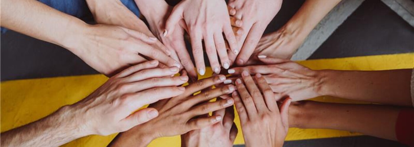 Mehrere Hände von Personen mit unterschiedlicher Hautfarbe und Geschlechtsidentität liegen übereinander in einem Kreis auf einem grauen Boden mit gelber Markierung; das Bild symbolisiert Vielfalt, Solidarität und Teamgeist.