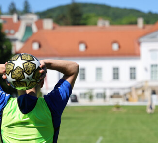 Eine Person im blauen Trikot und grünen Trainingsleibchen hält einen Fußball über dem Kopf zum Einwurf. Im Hintergrund sind eine Wiese, ein großes helles Gebäude mit rotem Dach, das Schloss Miller-Aichholz, und bewaldete Hügel zu sehen.