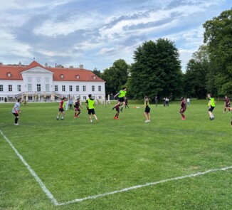Fußballszene direkt vor dem Tor mit mehreren Spieler:innen in grünen und weinroten Trikots. Eine Person springt zum Kopfball. Im Hintergrund ein historisches Gebäude mit rotem Dach und mehrere Zuschauer:innen auf der Wiese.