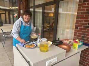 Eine Mitarbeiter:in steht im Außenbereich vor einem Tisch mit Kochutensilien und bereitet Speisen zu. Auf dem Tisch befinden sich eine Pfanne, Öl, Teig und Zutaten. Die Person richtet gerade ein frisch gebratenes Gericht auf einem Teller an. Im Hintergrund ist der Eingangsbereich des Hauses zu sehen.