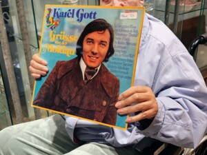 Eine ältere Person sitzt im Rollstuhl vor einer Glasvitrine mit Geschirr und hält eine Schallplatte mit dem Titel "Grüsse von der Moldau" von Karel Gott in den Händen. Die Person blickt direkt in die Kamera. Die Umgebung wirkt wie ein Verkaufsraum für gebrauchte Gegenstände.