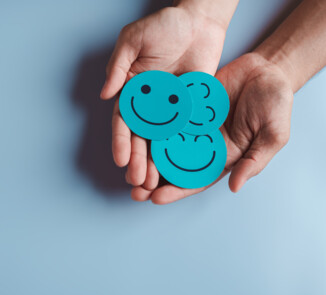 Symbolbild. Zwei geöffnete Hände halten drei türkisfarbene runde Karten mit verschiedenen Smileys, die Freude und positive Emotionen ausdrücken. Der Hintergrund ist einfarbig hellblau.