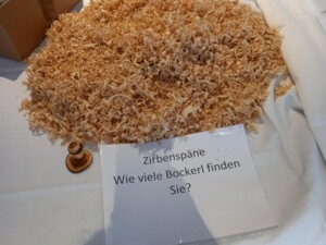 Ein Tisch mit einem großen Haufen Zirbenspäne. Daneben liegt ein kleines Holzobjekt und ein laminiertes Schild mit der Aufschrift "Zirbenspäne - Wie viele Bockerl finden Sie?". Die Szene lädt zum spielerischen Suchen ein.