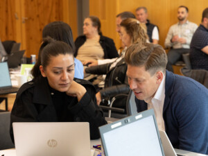 Zwei Personen sitzen nebeneinander vor einem Laptop, schauen konzentriert auf den Bildschirm und diskutieren etwas.